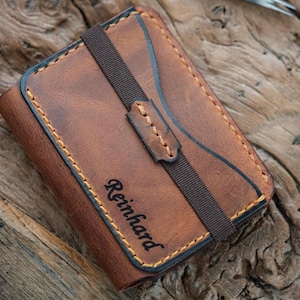 May include: A brown leather wallet featuring a dark brown elastic strap across the center and visible tan stitching along the edges. The name Reinhard is embossed in black script on the front surface of the leather wallet.