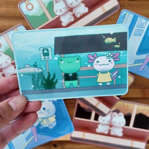 K&ouml;nnte beinhalten: Eine Sammlung farbenfroher Aufkleber mit niedlichen Cartoon-Charakteren. Ein Aufkleber zeigt einen Frosch im Anzug und einen Axolotl in einem gelben Kleid. Andere Aufkleber zeigen Hasen und Busszenen. Die Aufkleber sind rechteckig mit abgerundeten Ecken.