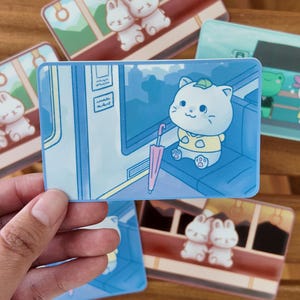 K&ouml;nnte beinhalten: Eine Sammlung farbenfroher Aufkleber mit Cartoon-Tieren. Ein Aufkleber zeigt eine wei&szlig;e Katze mit gr&uuml;nem Hut und rosa Regenschirm. Weitere Aufkleber zeigen Hasen und einen Frosch. Die Aufkleber haben eine gl&auml;nzende Oberfl&auml;che.