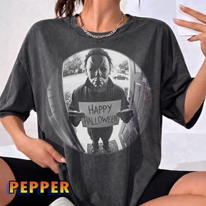Puede incluir: Una camiseta gris carbón de manga corta con un estampado gráfico circular de una figura enmascarada en una puerta, sosteniendo un cartel que dice Happy Halloween. El diseño imita una vista de ojo de pez de una calle suburbana.