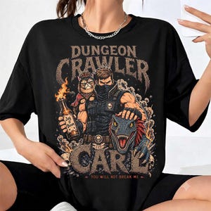 May include: A black t-shirt featuring a graphic print with the text "DUNGEON CRAWLER CARL" and "YOU WILL NOT BREAK ME". The design depicts a masked character, a cat wearing a crown, a dinosaur, and a bottle labelled "JUG O BOOM" with flames.