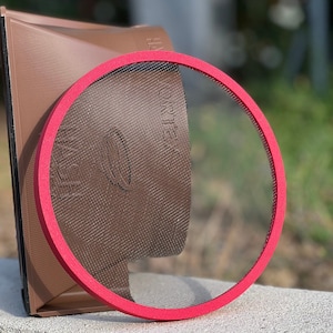 Può includere: Un colino rotondo a rete rossa con una base rettangolare marrone. Il colino ha una rete nera e la base ha le parole "HAY" e "CRIE" stampate su di essa.