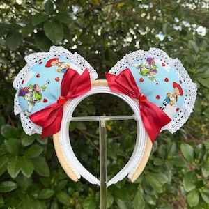 May include: A white headband with two large, round ears featuring a light blue fabric with cartoon characters, red hearts, and white lace trim. Each ear has a large red bow. The headband is cream-colored.