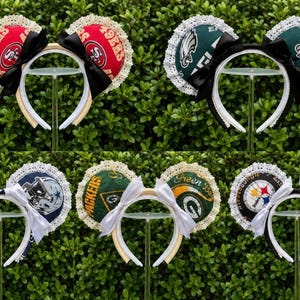 May include: A collection of six headbands featuring circular ears decorated with various NFL team logos, including the 49ers, Eagles, Cowboys, Packers, and Steelers. Each pair of ears is trimmed with white lace and accented with a solid color bow.
