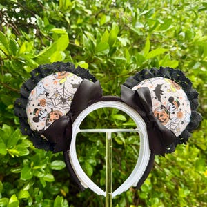 May include: A headband featuring two circular ears with a white fabric cover printed with Halloween motifs including ghosts, pumpkins, and spiderwebs. Each ear is trimmed with black lace and accented with a black satin bow on the front.