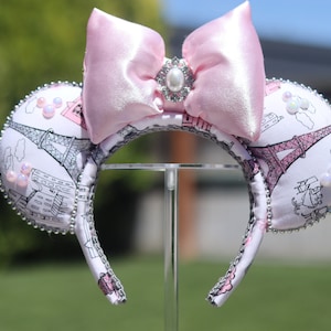 Könnte beinhalten: Rosa und weiße Minnie Mouse Ohren mit einer rosa Schleife und einer Perlen-Brosche. Die Ohren sind mit einem Schwarz-Weiß-Druck des Eiffelturms und anderer Pariser Sehenswürdigkeiten verziert.