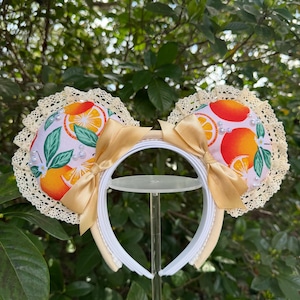 Könnte beinhalten: Ein Haarreif mit zwei runden Ohren aus weißem Stoff, bedruckt mit Orangen, grünen Blättern und weißen Blüten. Die Ohren sind mit cremefarbener Spitze eingefasst und mit kleinen weißen Perlen verziert. Zwei goldene Satinschleifen sind angebracht.