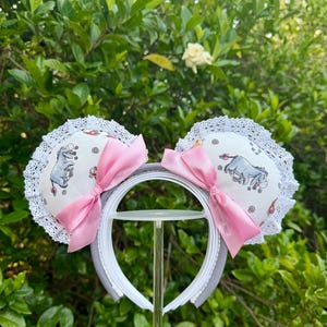 Puede incluir: Una diadema con dos orejas redondas cubiertas de tela blanca con un estampado repetitivo del personaje Igor. Cada oreja está adornada con encaje blanco y un lazo de satén rosa. La estructura de la diadema es de color gris claro y lisa.