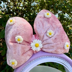 May include: A pink velvet headband with a large bow adorned with small white and yellow daisy flowers. The headband has a lavender band and a clear plastic piece.