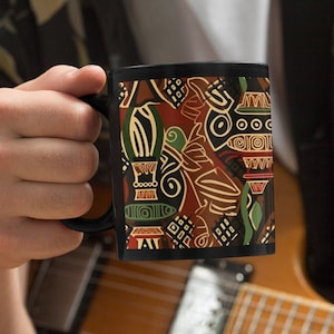 May include: A black coffee mug with a colorful geometric pattern. The mug is being held by a person who is sitting with a guitar.
