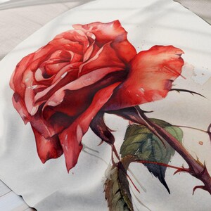 May include: A white fleece throw blanket featuring a watercolour painting of a single red rose with green leaves and thorns. The rose is positioned in the centre of the blanket and faces the right side of the image. The blanket is displayed on a bed with grey pillows.