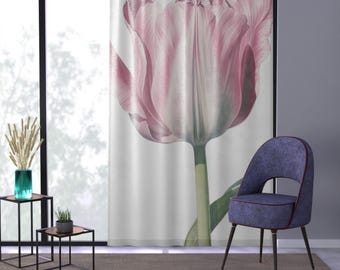 Op de afbeelding: Een wit gordijn met een grote roze tulp erop gedrukt. De tulp is scherp en het gordijn is licht wazig. Een blauwe stoel met zwarte poten staat op de voorgrond.