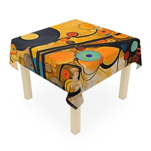 May include: A square tablecloth with a colorful abstract design featuring circles, lines, and swirls in shades of yellow, orange, red, blue, black, and white. The tablecloth is draped over a wooden table with four legs.