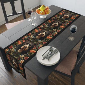 Puede incluir: Un camino de mesa negro y blanco con detalles florales. El camino de mesa está sobre una mesa de madera oscura con un tazón de manzanas, copas de vino y cubiertos.