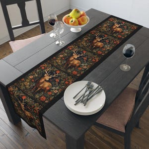 May include: A black table runner with a tapestry design featuring a repeating pattern of deer in a forest setting. The runner is laid on a dark wood table with a set of white plates and cutlery, two wine glasses, and a bowl of apples.