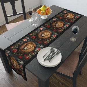 May include: A black table runner with a repeating pattern of lion faces surrounded by colorful flowers. The runner is laid out on a dark wood table with a bowl of apples and two wine glasses.