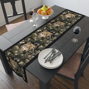 May include: A black table runner with a repeating pattern of wolf faces surrounded by green foliage and white flowers. The runner is laid out on a dark wood table with a set of white plates and silverware.