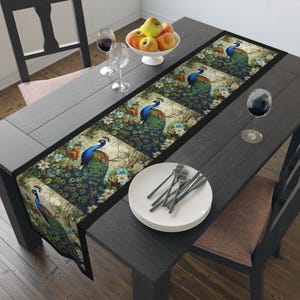 May include: A black and green table runner with a peacock design. The runner is on a dark wood table with a bowl of apples, two wine glasses, and a plate with silverware.