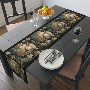 May include: A black table runner with a repeating pattern of white sheep faces surrounded by colorful flowers. The runner is on a dark wood table with a plate and silverware.
