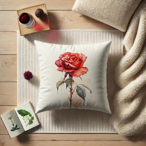May include: A white throw pillow with a watercolor painting of a single red rose with green leaves and a stem with thorns. The pillow is on a white knitted surface.