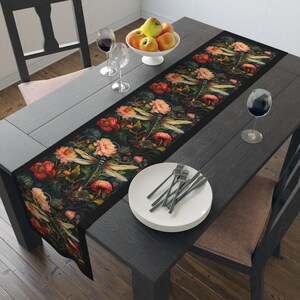 May include: A black table runner with a floral and dragonfly design. The runner features pink, red, and green flowers with blue dragonflies. The runner is on a black wooden table with a white plate and silverware.