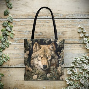 May include: Tote bag featuring a detailed illustration of a wolf's face surrounded by foliage. The wolf has light brown fur and yellow eyes. The bag has a black handle and a dark background.