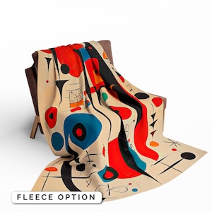 Pode incluir: Uma manta de lã polar com um design abstrato que apresenta formas pretas, vermelhas, azuis e amarelas sobre um fundo creme. A manta está dobrada e drapeada sobre uma cadeira.
