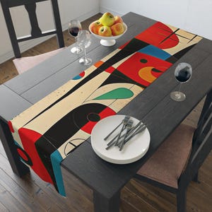 May include: A colorful abstract table runner with geometric shapes in red, blue, yellow, green, and black on a cream background. The runner is placed on a dark gray table with a white plate and silverware.