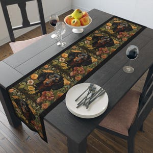 May include: A black and tan dachshund table runner with a floral pattern. The runner is on a dark wood table with two chairs and a bowl of apples.