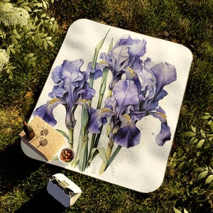 May include: A white picnic blanket with a watercolor illustration of purple irises. The flowers are in full bloom and have a delicate, hand-painted look.