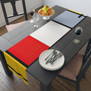 Puede incluir: Un camino de mesa con un diseño geométrico con rayas amarillas, negras, rojas, blancas y grises. El camino de mesa está colocado sobre una mesa de madera oscura con un cuenco de manzanas y dos copas de vino.