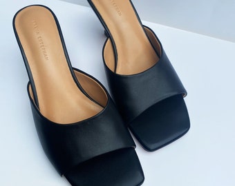 May include: A pair of black leather heeled mules with a square toe and open back. The shoes have a low heel and are labeled "Theya Estherman".