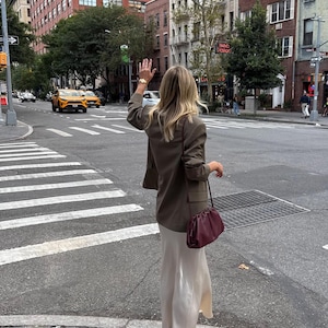 May include: An olive green blazer with long sleeves and a structured collar, paired with a floor-length cream-colored satin skirt. A dark burgundy leather clutch bag with a gathered top and a thin shoulder strap is worn over the shoulder.