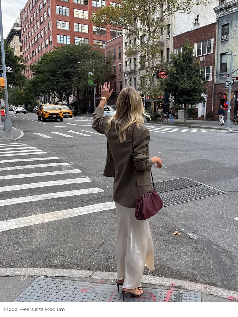 May include: An olive green blazer with long sleeves and a structured collar, paired with a floor-length cream-colored satin skirt. A dark burgundy leather clutch bag with a gathered top and a thin shoulder strap is worn over the shoulder.