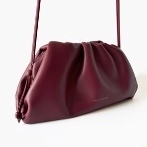 May include: A deep burgundy, ruched pouch bag with a soft, gathered texture and a thin shoulder strap. The front of the bag features the gold-toned text FREYA ESTEPHAN printed in a small, centered font. The design includes a drawstring-style closure.