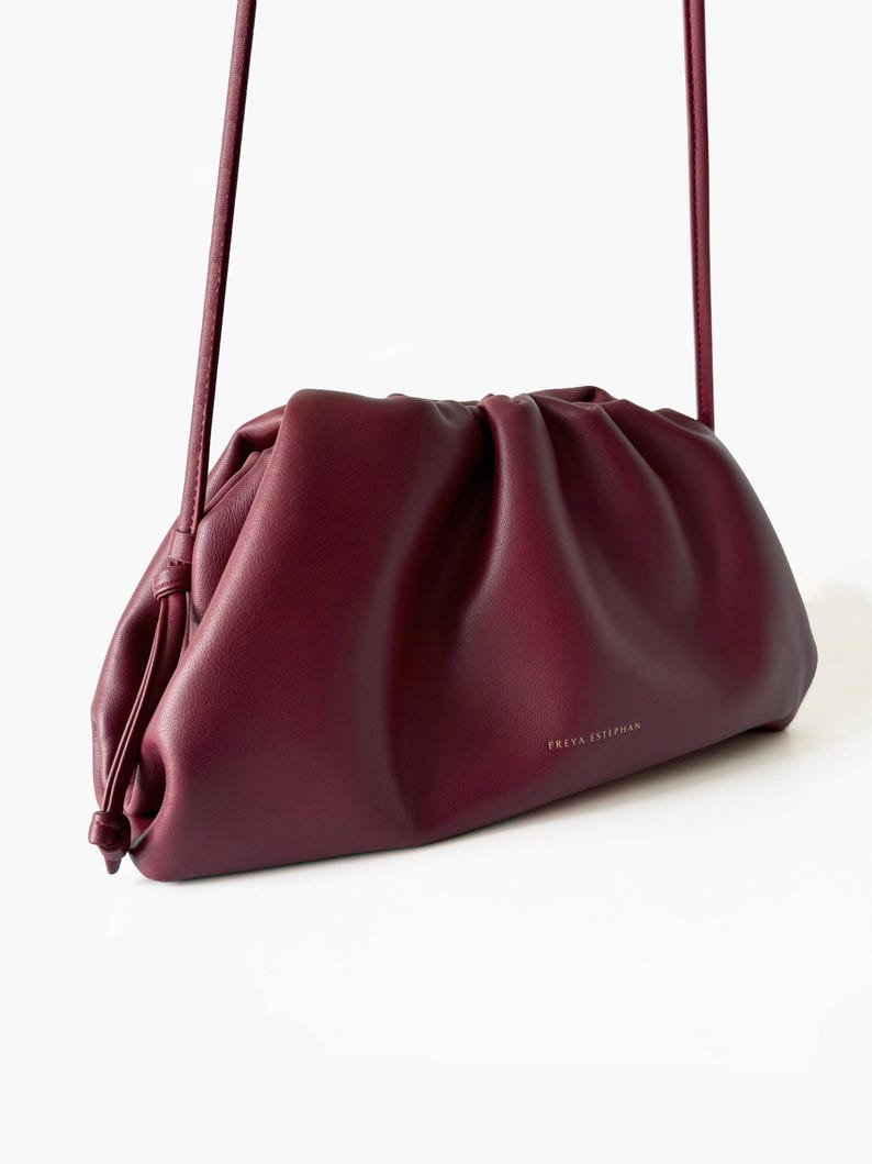 May include: A deep burgundy, ruched pouch bag with a soft, gathered texture and a thin shoulder strap. The front of the bag features the gold-toned text FREYA ESTEPHAN printed in a small, centered font. The design includes a drawstring-style closure.