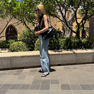Puede incluir: Una persona con una camiseta negra, vaqueros azul claro y sandalias negras, lleva un bolso negro con una correa de cadena. El fondo presenta un edificio, &aacute;rboles y un cielo soleado.