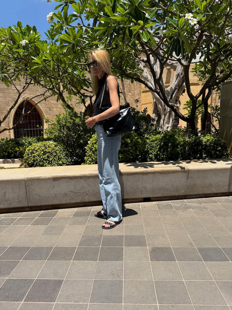 Puede incluir: Una persona con una camiseta negra, vaqueros azul claro y sandalias negras, lleva un bolso negro con una correa de cadena. El fondo presenta un edificio, &aacute;rboles y un cielo soleado.