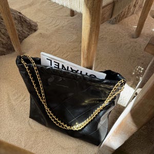 Puede incluir: Bolso tote de cuero negro acolchado con una correa de cadena dorada. La bolsa est&aacute; llena de una revista. La bolsa est&aacute; sobre una superficie arenosa, con patas de silla de madera en el fondo.