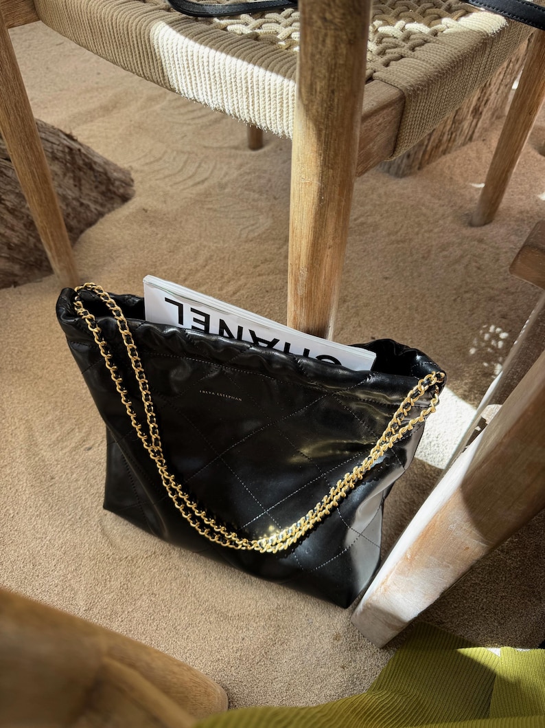 Puede incluir: Bolso tote de cuero negro acolchado con una correa de cadena dorada. La bolsa est&aacute; llena de una revista. La bolsa est&aacute; sobre una superficie arenosa, con patas de silla de madera en el fondo.