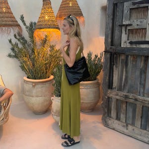 Puede incluir: Una mujer con un vestido largo verde oliva y sandalias negras, llevando un bolso negro. El fondo muestra grandes macetas, l&aacute;mparas colgantes tejidas y una puerta de madera r&uacute;stica.