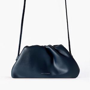 May include: A dark navy blue clutch bag with a soft, gathered, ruched top design and a long, thin shoulder strap. The front of the bag features the gold-toned text FREYA ESTEPHAN centered near the bottom edge, showcasing a smooth, matte finish.