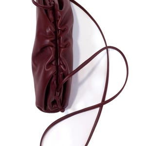 May include: A deep burgundy leather pouch bag featuring a gathered, ruched top closure and a thin, matching leather shoulder strap. The bag has a soft, rounded silhouette with visible folds in the leather material, creating a textured appearance.