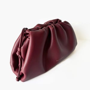 May include: A deep burgundy, soft-structured clutch bag with a gathered, ruched top opening and a thin, knotted side drawstring detail. The bag features a smooth, matte finish and the gold-toned text "FREYA COLLECTION" printed on the front.