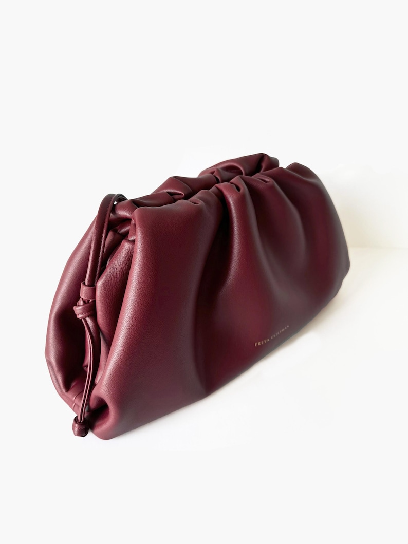 May include: A deep burgundy, soft-structured clutch bag with a gathered, ruched top opening and a thin, knotted side drawstring detail. The bag features a smooth, matte finish and the gold-toned text "FREYA COLLECTION" printed on the front.