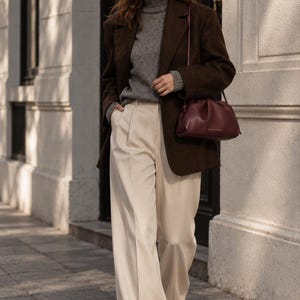 May include: A dark brown wool blazer worn over a grey turtleneck sweater, paired with cream-colored wide-leg trousers. A deep burgundy leather clutch bag with a shoulder strap is carried, and dark-tinted sunglasses are worn on the face.