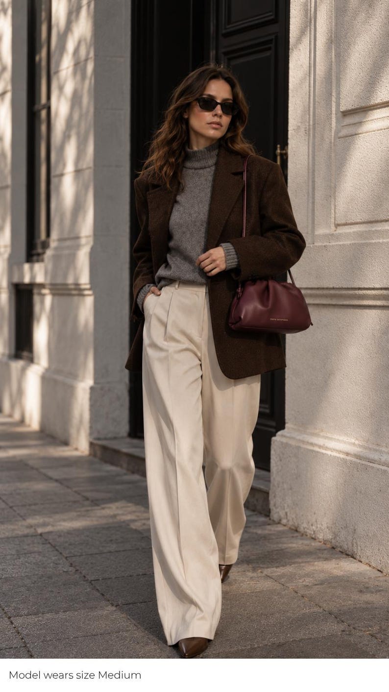 May include: A dark brown wool blazer worn over a grey turtleneck sweater, paired with cream-colored wide-leg trousers. A deep burgundy leather clutch bag with a shoulder strap is carried, and dark-tinted sunglasses are worn on the face.