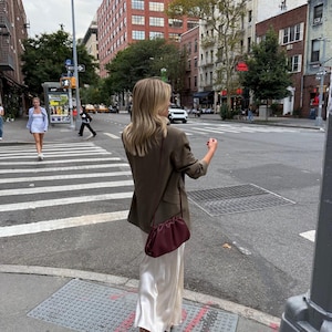 May include: A brown blazer worn over a long, cream-colored satin skirt. A dark burgundy, ruched, pouch-style shoulder bag with a thin strap is worn across the body. The outfit is paired with dark-colored heels on a city street crosswalk.