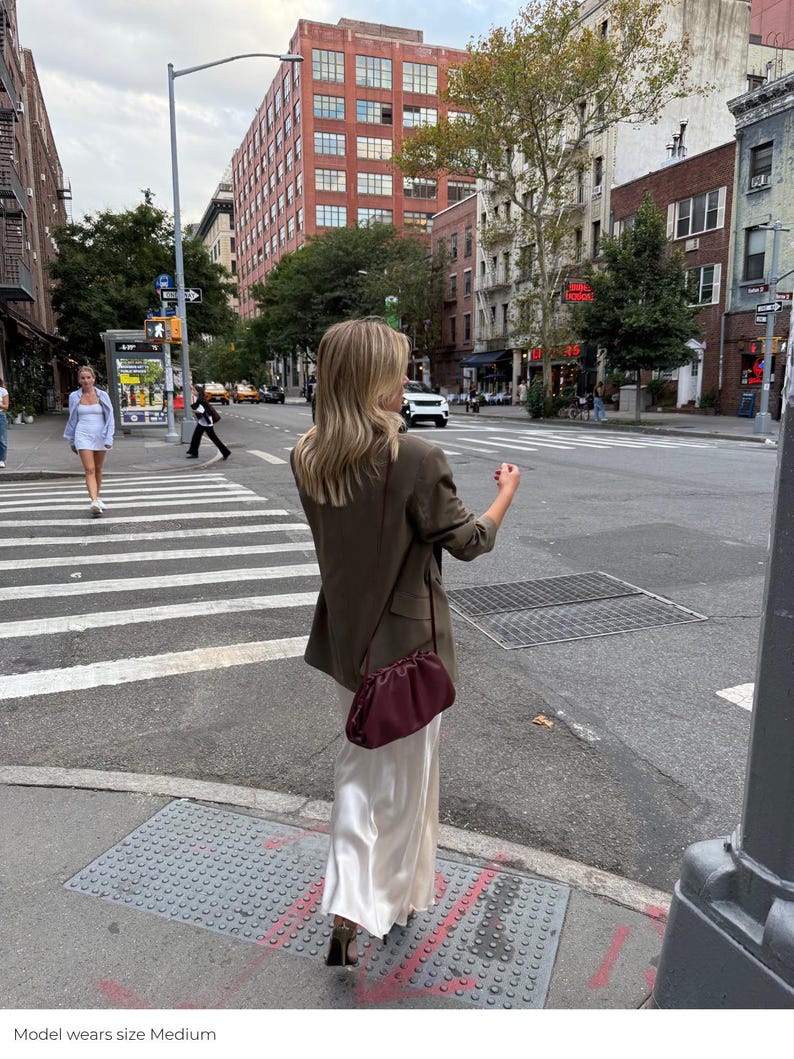 May include: A brown blazer worn over a long, cream-colored satin skirt. A dark burgundy, ruched, pouch-style shoulder bag with a thin strap is worn across the body. The outfit is paired with dark-colored heels on a city street crosswalk.