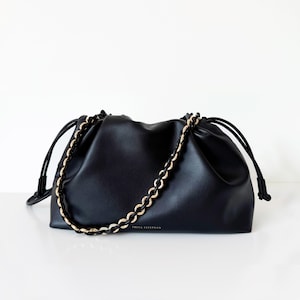 May include: A black handbag with a smooth finish, featuring a drawstring closure on both sides and a decorative shoulder strap composed of intertwined black and gold-tone chain links. The brand name FREYA ESTEPHAN is printed in small gold letters.